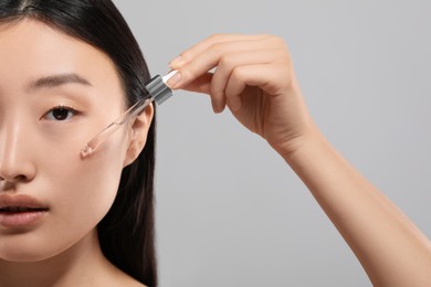 Beautiful young woman applying cosmetic serum onto her face on grey background, closeup