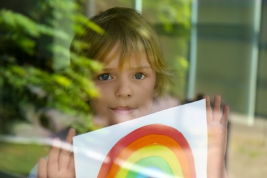 Photo of Little boy with picture of rainbow near window, view from outdoors. Stay at home concept