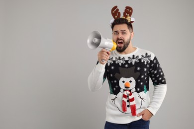 Photo of Young man in Christmas sweater and reindeer headband shouting in megaphone on grey background. Space for text