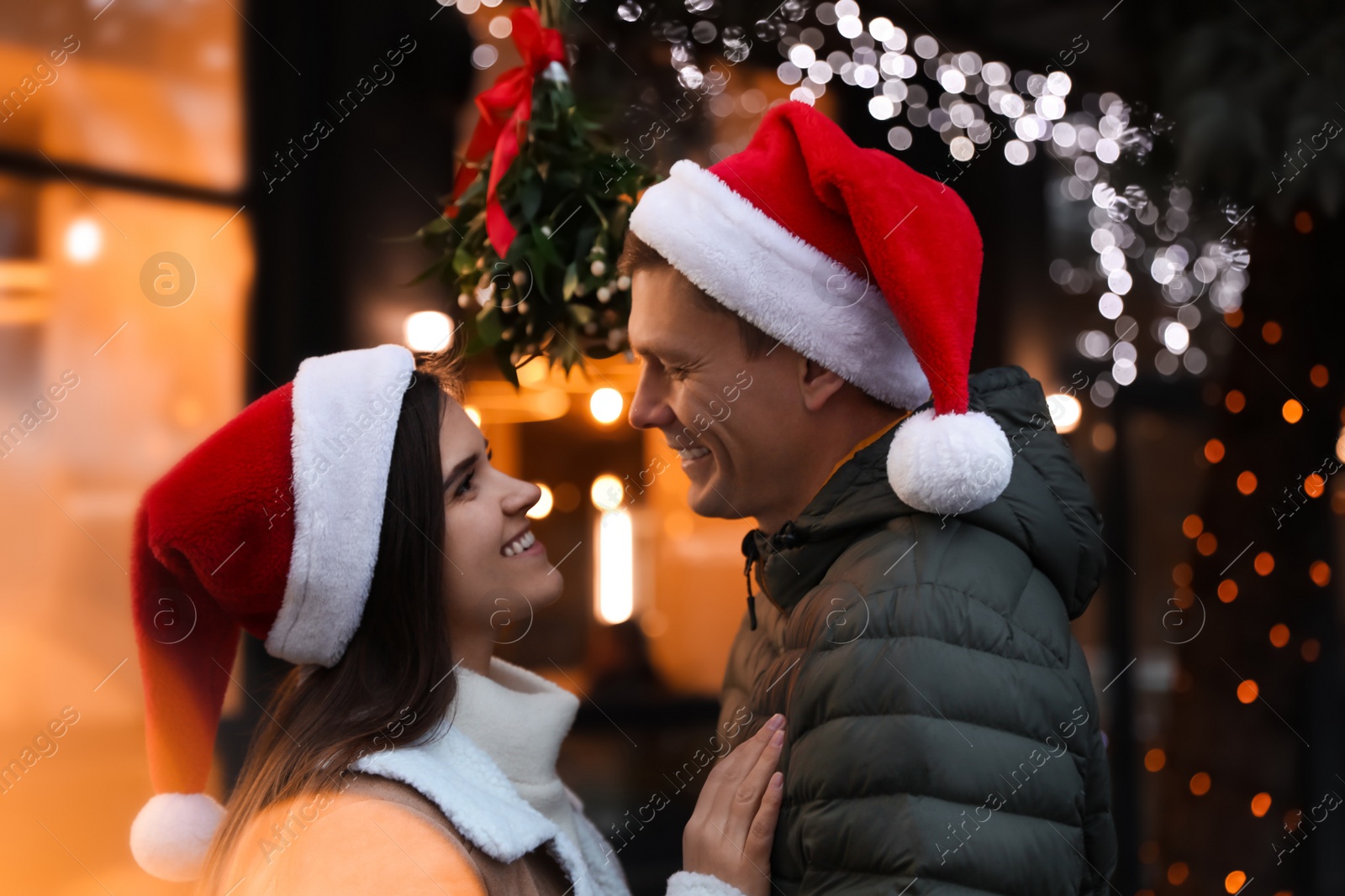 Photo of Happy couple in Santa hats standing under mistletoe bunch outdoors