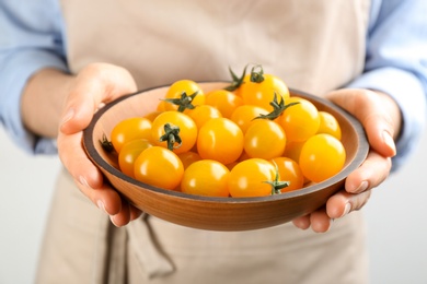 Photo of Woman holding wooden bowl of yellow tomatoes on light background, closeup