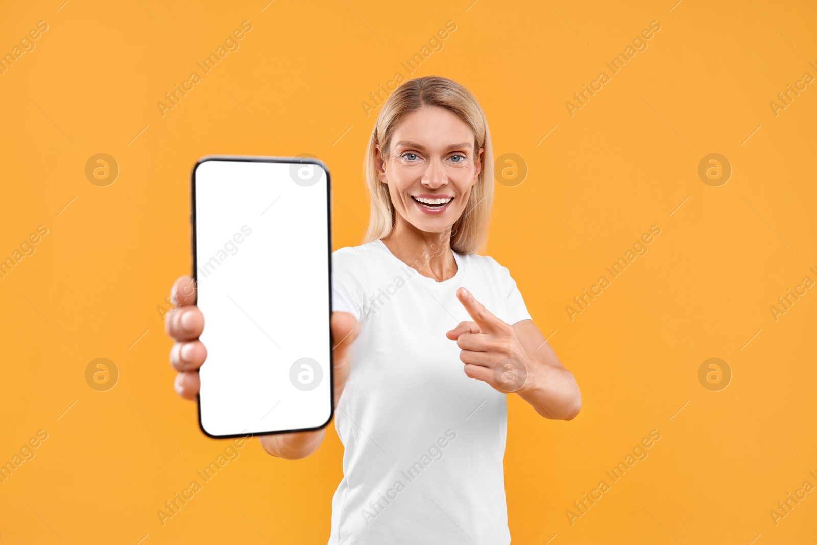 Photo of Happy woman holding smartphone and pointing at blank screen on orange background