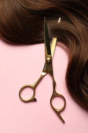 Photo of Professional hairdresser scissors with brown hair strand on pink background, top view