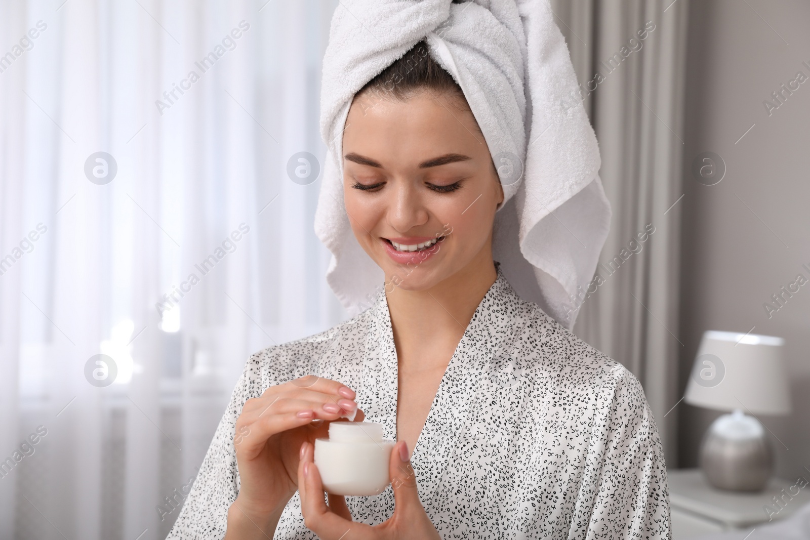 Photo of Beautiful young woman with hair wrapped in towel holding face cream at home