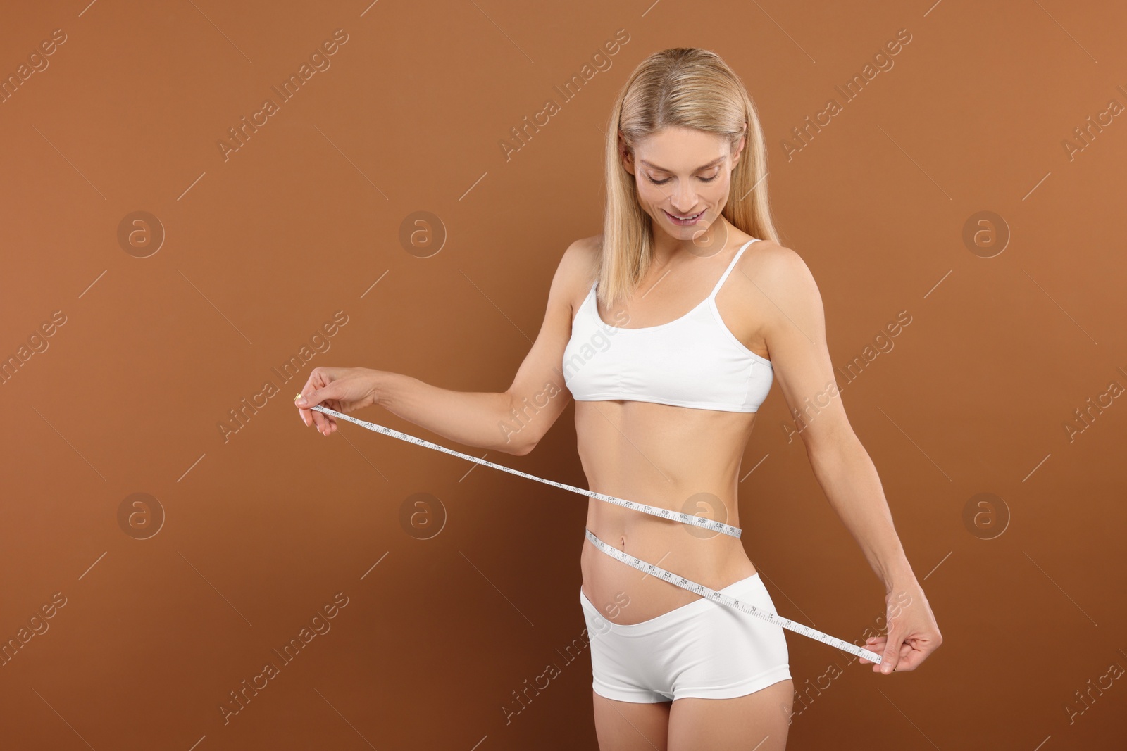 Photo of Slim woman measuring waist with tape on brown background, space for text. Weight loss