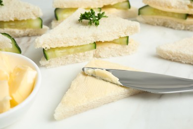 Photo of Spreading butter on tasty sandwiches with cucumber and parsley on white marble table, closeup