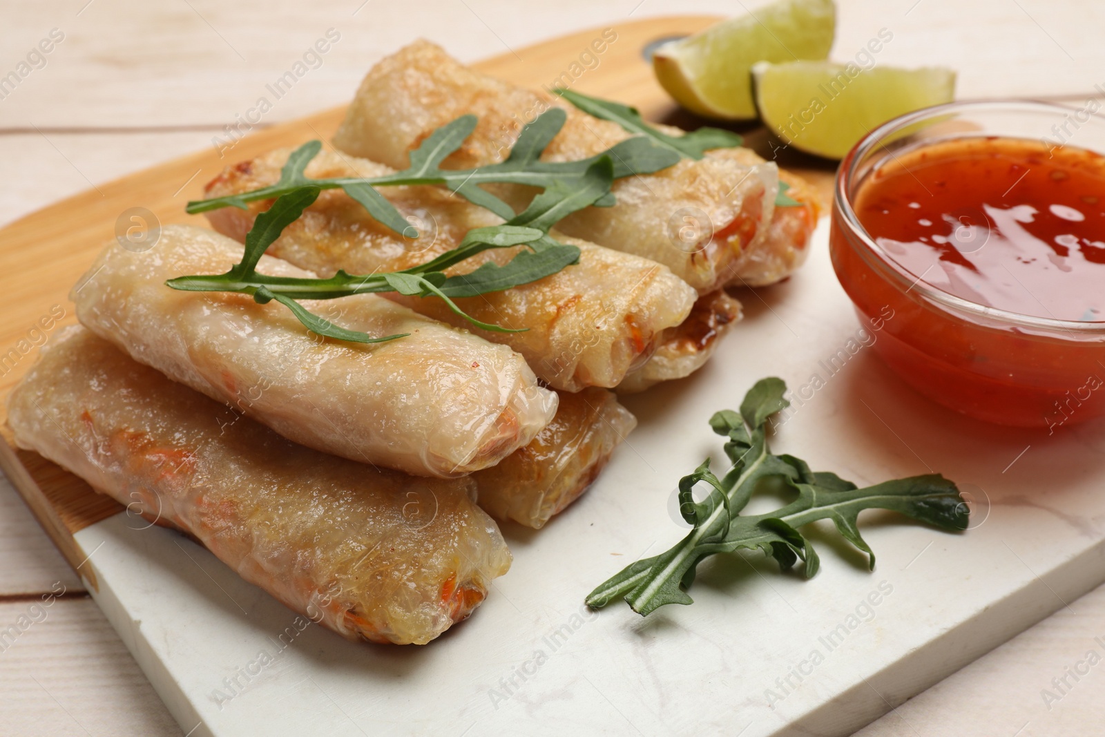 Photo of Tasty fried spring rolls, lime, arugula and sauce on light wooden table, closeup