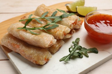 Photo of Tasty fried spring rolls, lime, arugula and sauce on light wooden table, closeup