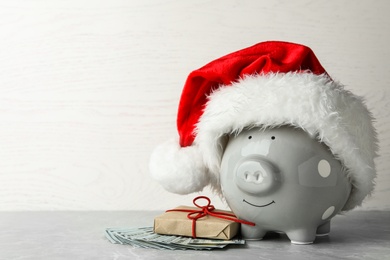 Photo of Piggy bank with Santa hat, gift box and dollar banknotes on grey table. Space for text