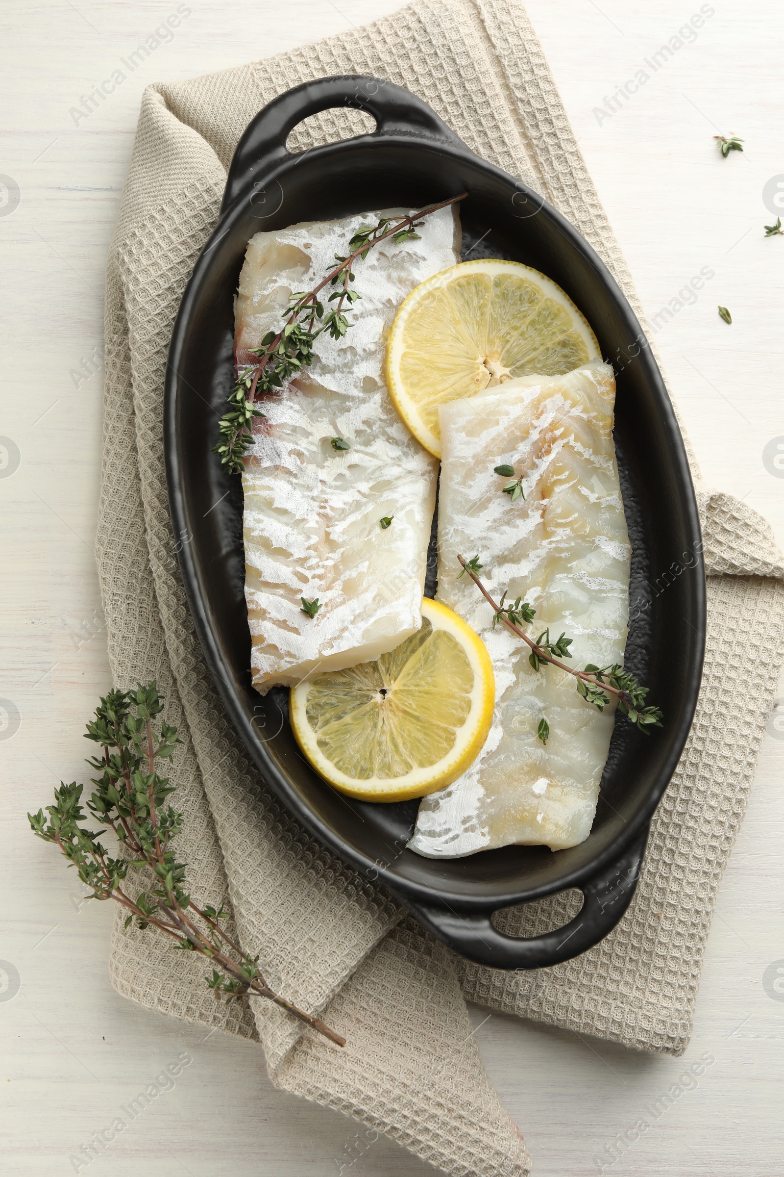 Photo of Fresh raw cod fillets with thyme and lemon in baking dish on white wooden table, top view