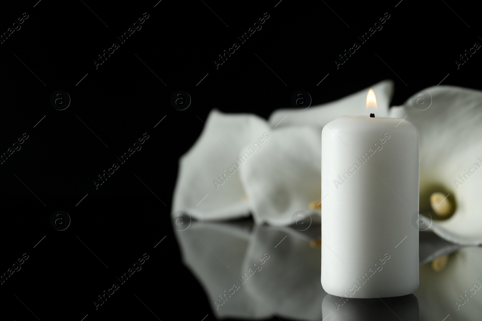 Photo of Burning candle and white calla lily flowers on black mirror surface in darkness, closeup with space for text. Funeral symbol