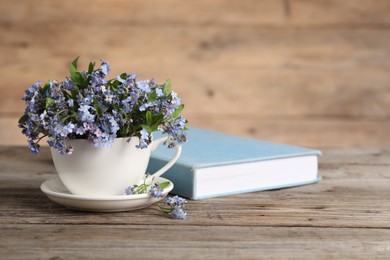 Beautiful forget-me-not flowers in cup, saucer and book on wooden table. Space for text