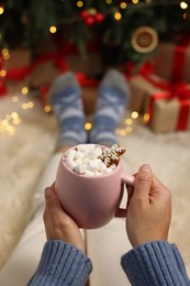 Photo of Woman holding cup of delicious cocoa with marshmallows near Christmas tree at home, closeup, Space for text
