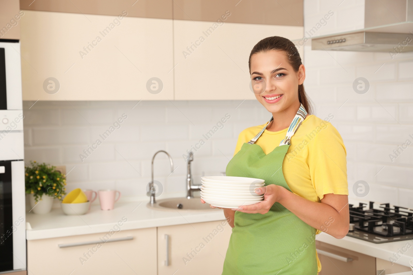 Photo of Beautiful young woman holding stack of clean dishes in kitchen