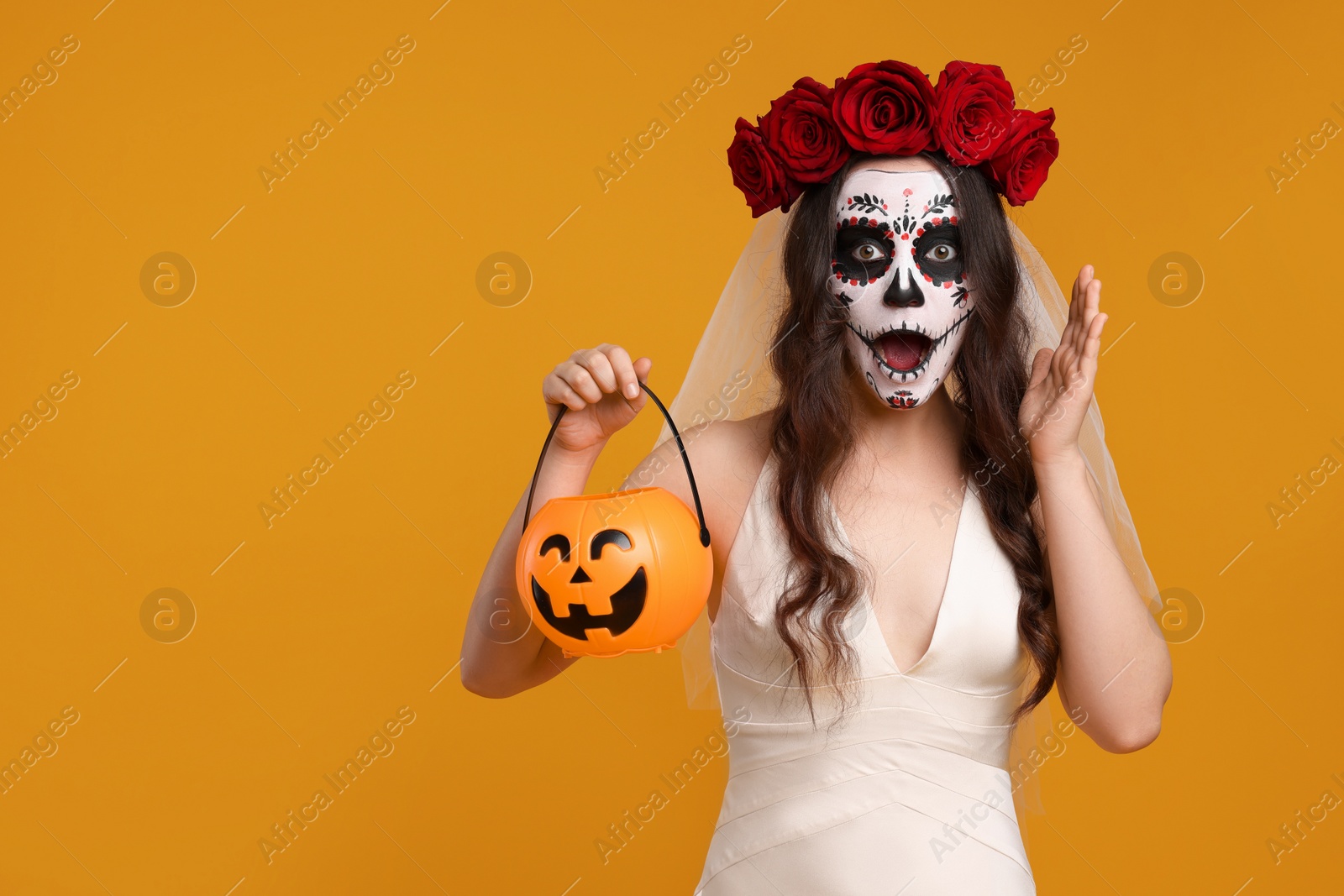 Photo of Emotional young woman in scary bride costume with sugar skull makeup, flower crown and pumpkin bucket on orange background, space for text. Halloween celebration