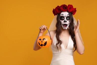 Photo of Emotional young woman in scary bride costume with sugar skull makeup, flower crown and pumpkin bucket on orange background, space for text. Halloween celebration