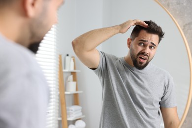 Photo of Emotional man examining his head near mirror in bathroom. Dandruff problem