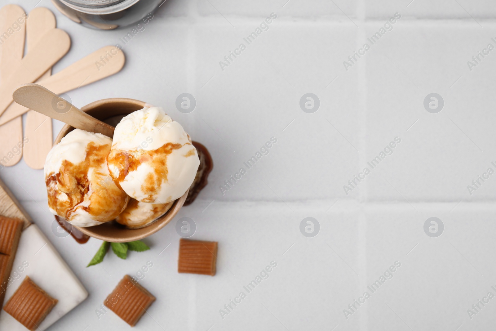 Photo of Scoops of ice cream with caramel sauce and candies on white tiled table, flat lay. Space for text