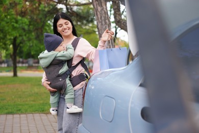 Mother holding her child in sling (baby carrier) while putting shopping bags into car trunk outdoors