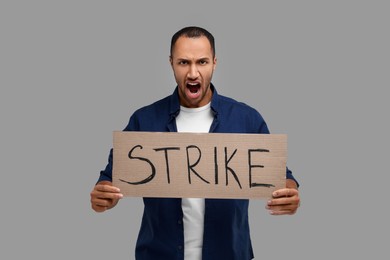 Angry man holding cardboard banner with word Strike on grey background