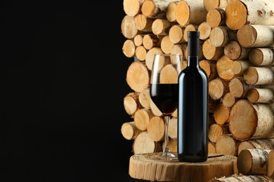 Stylish presentation of red wine in bottle and wineglass near wooden logs on black background, space for text