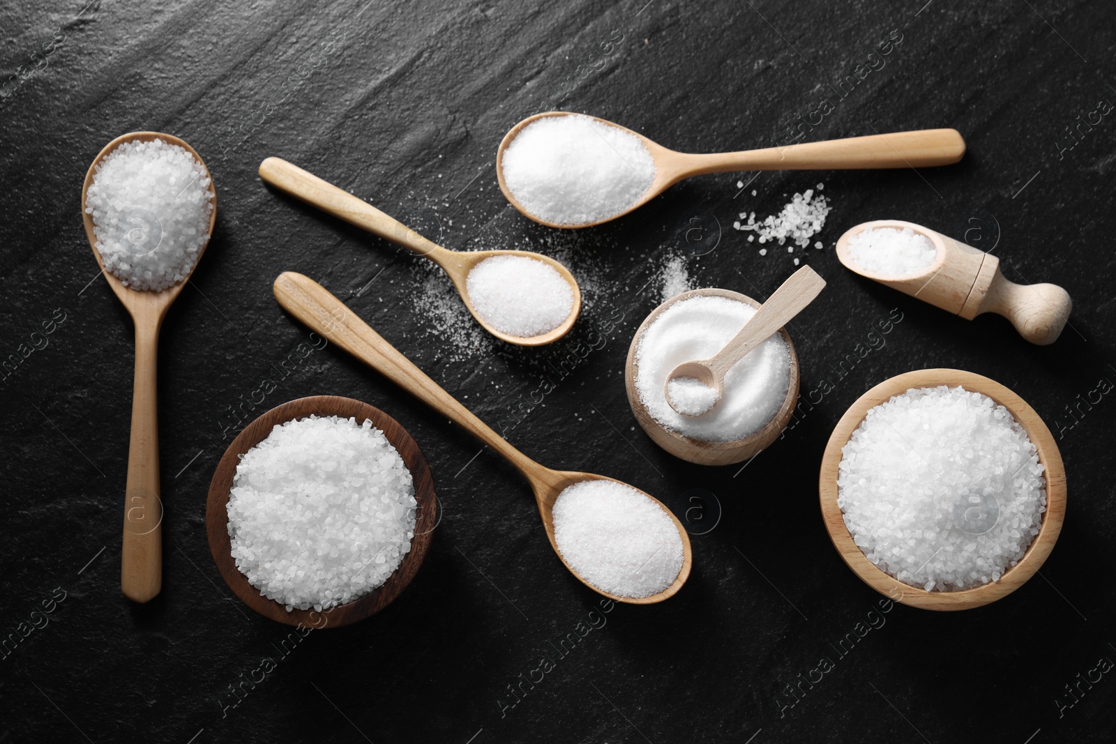 Photo of Organic white salt in bowls and spoons on black table, flat lay