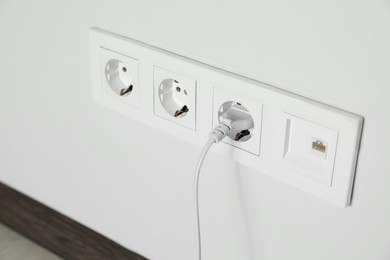 Photo of Power sockets with inserted plug on white wall indoors, closeup. Electrical supply