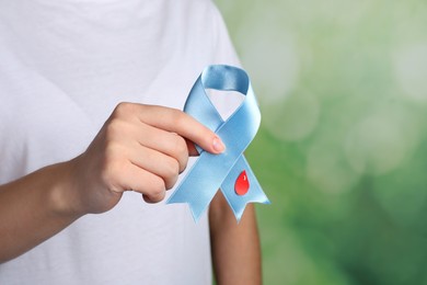 Photo of Woman holding light blue ribbon with paper blood drop against blurred green background, closeup. World Diabetes Day