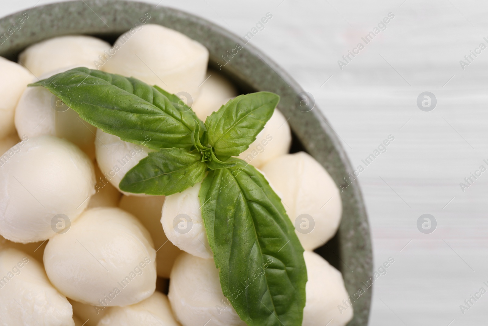 Photo of Tasty mozzarella balls and basil leaves in bowl on white wooden table, top view. Space for text