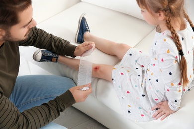 Father applying bandage on daughter's injured knee at home, closeup. First aid