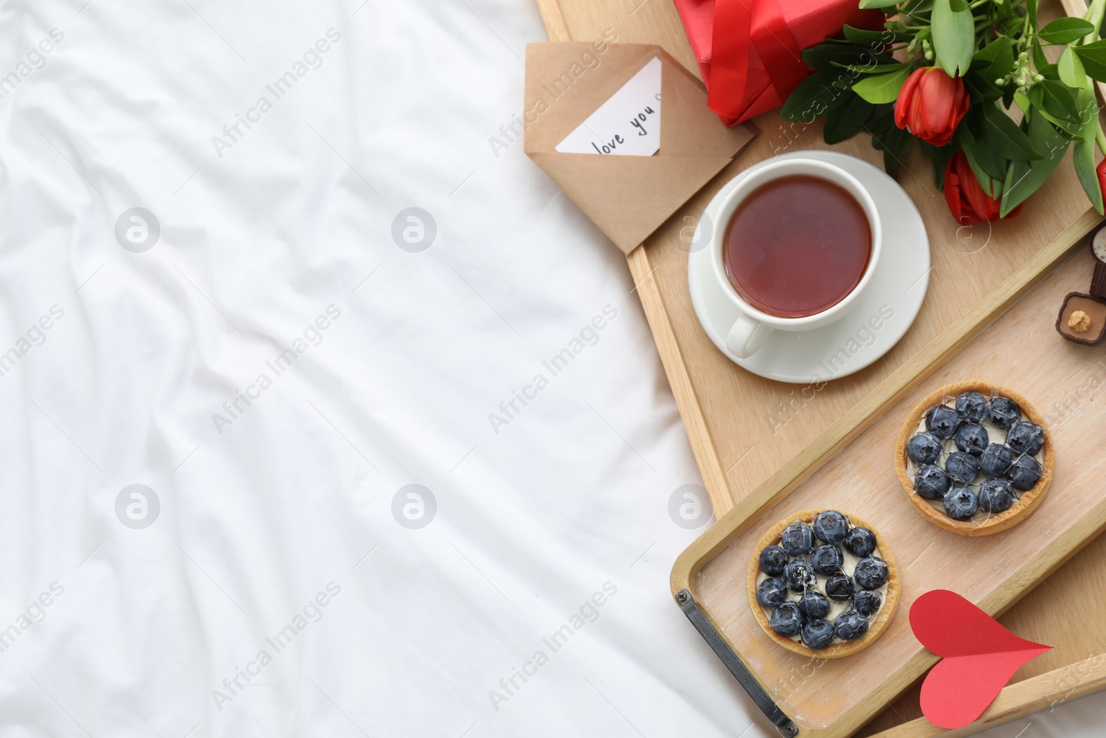 Photo of Tasty breakfast served in bed. Delicious tartlets, tea, flowers and card with phrase Love You on tray, top view. Space for text