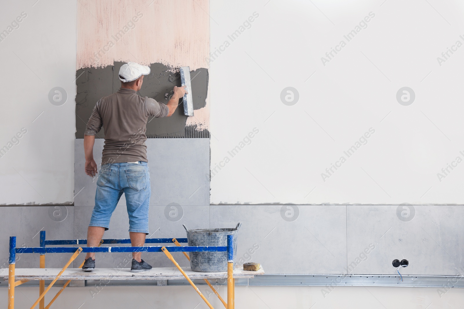 Photo of Worker spreading adhesive mix over wall for tile installation indoors, back view