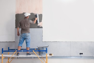 Worker spreading adhesive mix over wall for tile installation indoors, back view