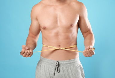 Young man with slim body using measuring tape on light blue background, closeup view