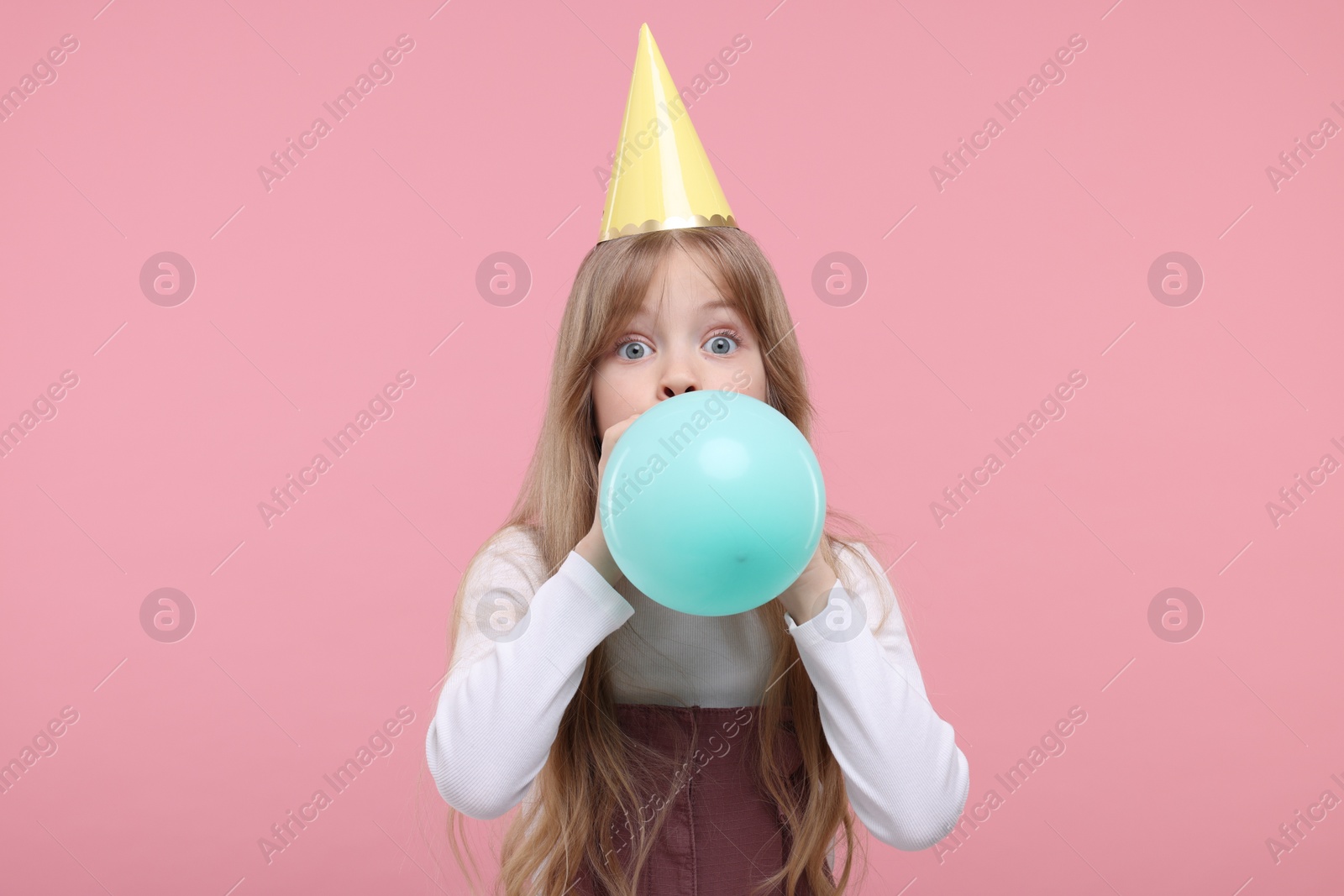 Photo of Cute little girl in party hat inflating balloon on pink background