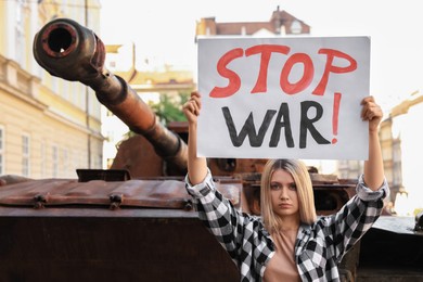 Photo of Sad woman holding poster with words Stop War near broken tank in city