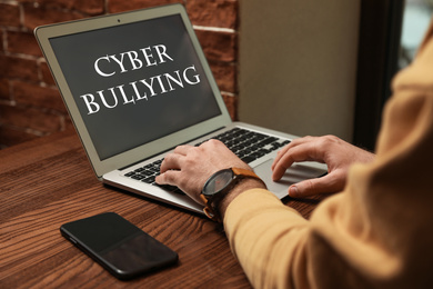 Young man using laptop at wooden table, closeup. Cyber bullying