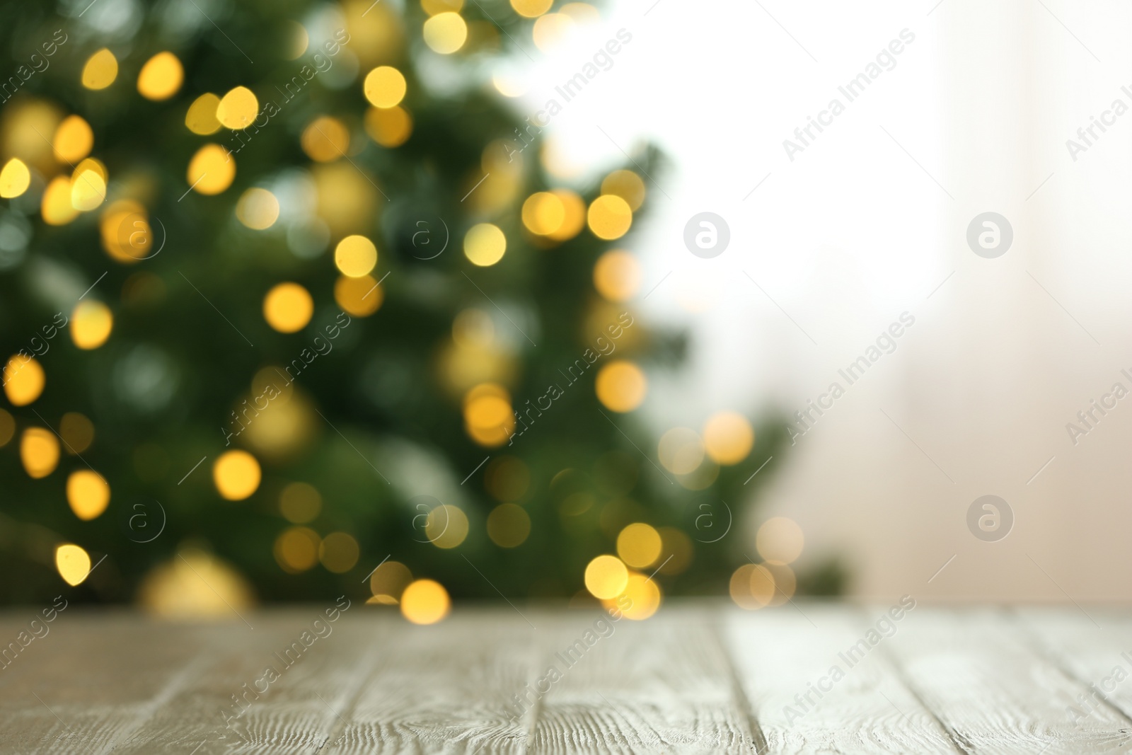 Photo of Blurred view of fir tree with glowing Christmas lights near table indoors. Festive mood