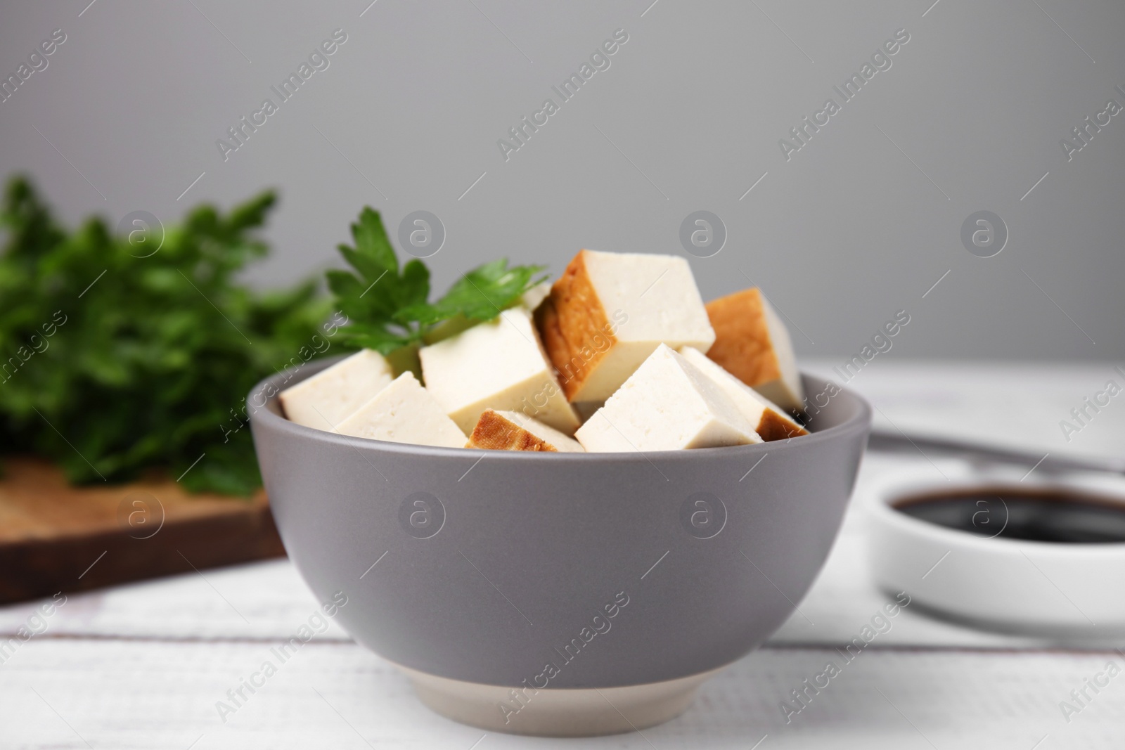 Photo of Bowl of smoked tofu cubes, soy sauce and parsley on white wooden table, closeup