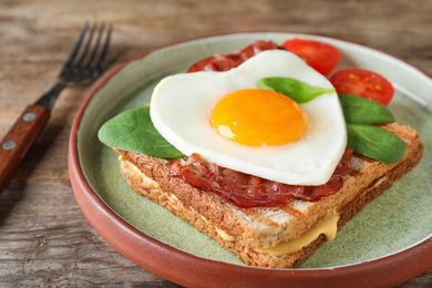 Plate of tasty sandwich with heart shaped fried egg and  bacon on wooden table, closeup