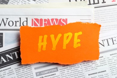 Image of Orange paper piece with word Hype on pile of newspapers, top view