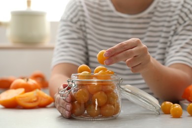 Photo of Woman putting yellow tomatoes into glass jar at light kitchen table, closeup. Pickling vegetables