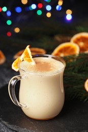 Glass cup of delicious eggnog with dried orange slice and fir branch on black table