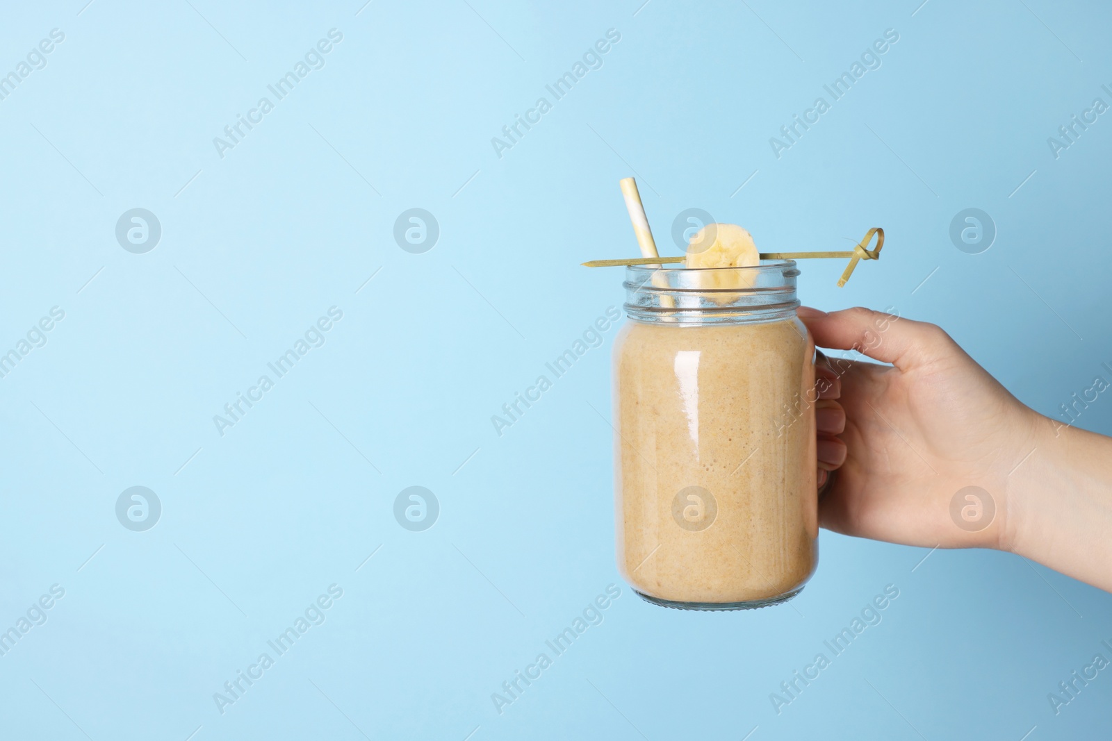 Photo of Woman holding mason jar with tasty smoothie, banana and straw on light blue background, closeup. Space for text