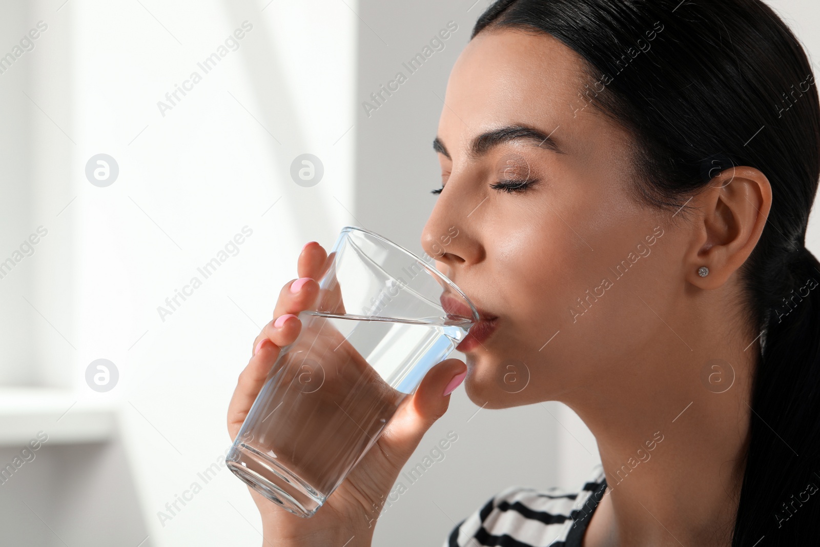 Photo of Beautiful young woman drinking water indoors, closeup. Refreshing drink