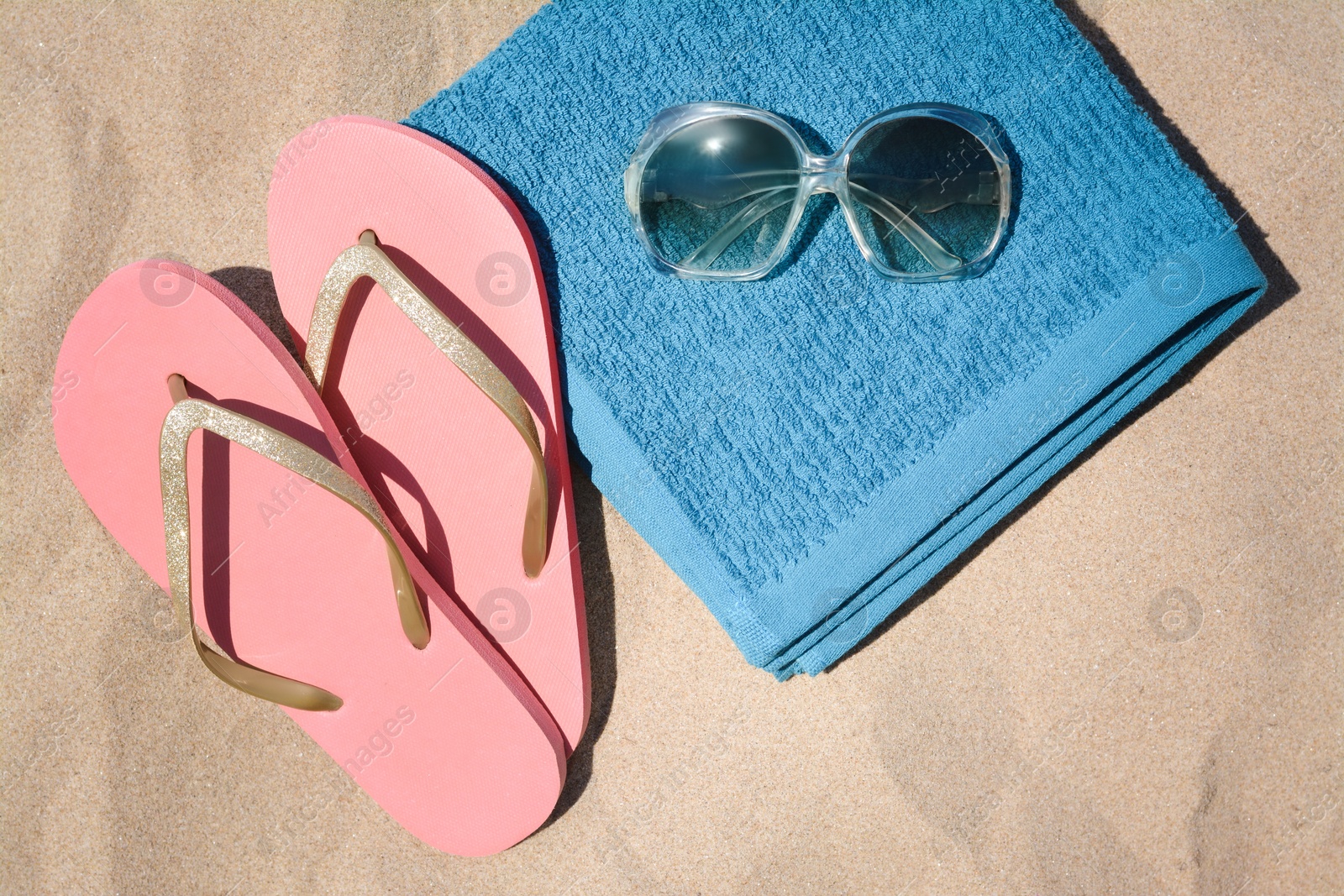 Photo of Folded soft blue beach towel with flip flops and sunglasses on sand, flat lay