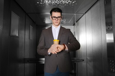 Photo of Young businessman with cup of hot drink checking time in elevator