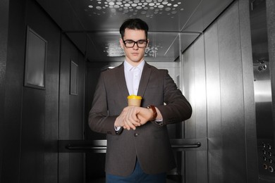 Young businessman with cup of hot drink checking time in elevator