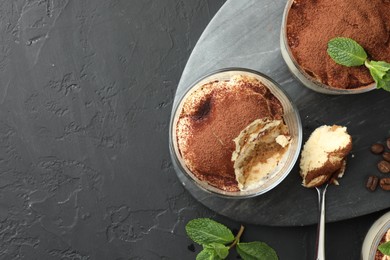 Delicious tiramisu in glasses, spoon, mint leaves and coffee beans on black table, top view. Space for text