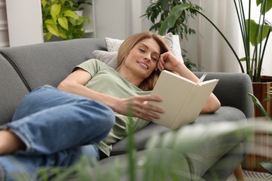 Photo of Woman reading book on sofa surrounded by beautiful potted houseplants at home